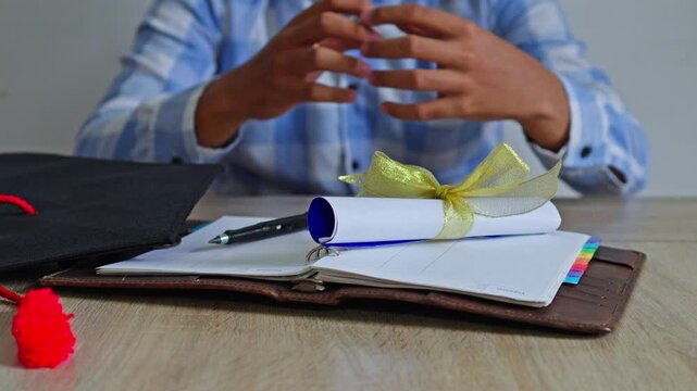Close-up of graduation scroll being set on wooden table with leather notebook and mortarboard
