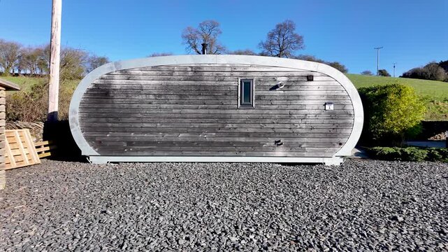A modern wooden glamping pod cabin on gravel set against rolling green hills and clear blue sky.