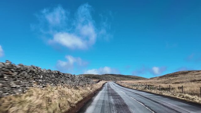 POV view from a car driving an empty country road through North Pennines moorland under a bright blue sky. slow motion