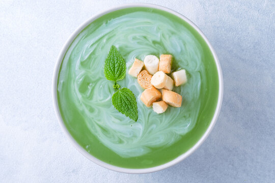 Close up of a bowl of mint or nettle leaves green soup sits on a blue background. The soup is swirled with cream, topped croutons. top view. Healthy food