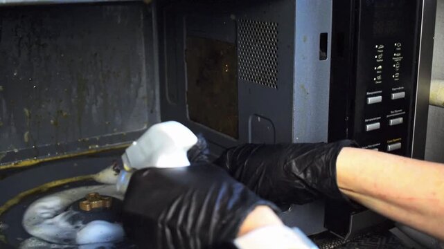 Hand in rubber glove cleaning very dirty microwave oven inside with yellow sponge and chemical spray, removing grease and food splatters. Kitchen equipment interior cleanup.