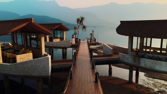 Overwater bungalows with a wooden pier stretch across a calm lake, reflecting the warm glow of sunset and providing a serene luxury resort experience with mountains in the background