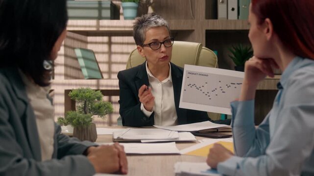 Caucasian senior mature business woman startup owner showing graphs to multicultural women workers businesswomen boss consulting clients females demonstrate document with data discuss marketing result