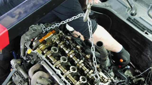 Technician works under hood of car near hanging engine. Technician returns system components to original condition repairing damage