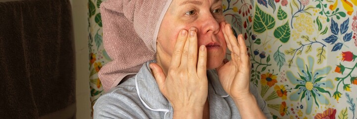 A 50-year-old woman prepares her skin by applying cream in her bathroom. She wears a towel on her head and stands in front of a colorful shower curtain during her morning routine, banner © pundapanda