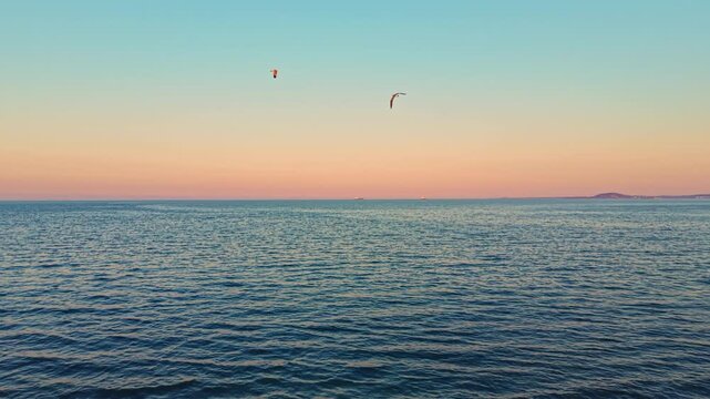 The sky shows warm colors as the sun sets over the ocean. Waves gently move in the water. A distant landmass is visible against the horizon. Birds fly in the air above the scene.