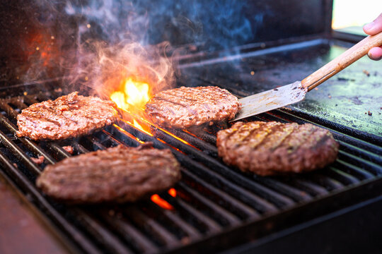 Hamburguesas de ternera cocin&aacute;ndose en una barbacoa con llamas y humo.