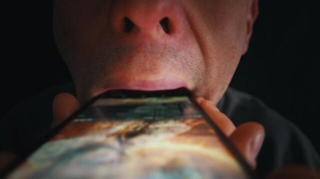 A man holds a glowing smartphone up to his wide open mouth as if trying to eat the screen. The dark room emphasizes the bright light from the device illuminating his face.