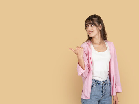 Smiling young woman pointing thumb over shoulder with confident expression, recommending or indicating direction concept image. Isolated on beige background with copy space for advertising use.