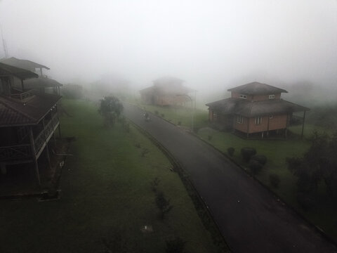 Aerial view of modern buildings, dark roofs, warm walls, veiled by dense fog, contrasting winding road, green slopes, Obudu, Cross River, Nigeria.