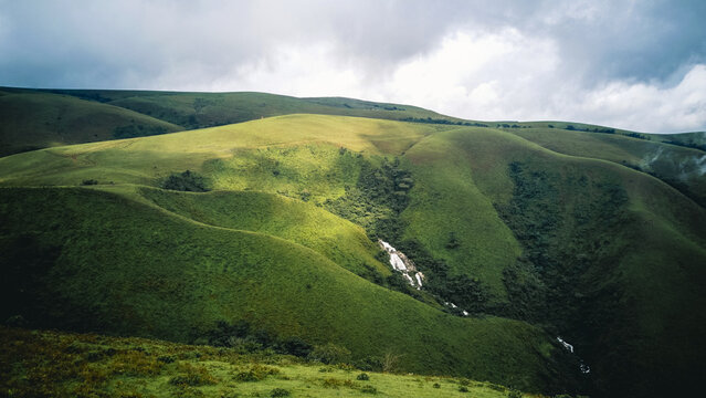 Aerial view of lush emerald slopes bathed in a patchwork of light and shadow, featuring a cascading silver waterfall under a dramatic cloudy sky, Obudu, Cross River, Nigeria.