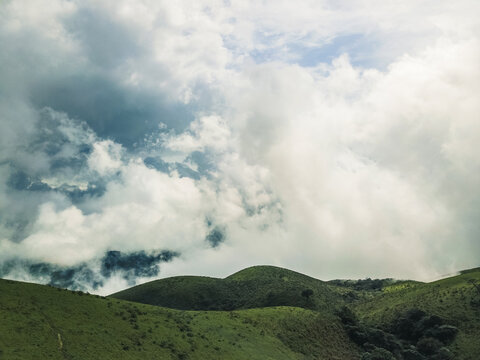 Aerial view of rolling green hills shrouded in a dramatic interplay of bright white and deep grey clouds under a peek of blue sky, Obudu, Cross River, Nigeria.