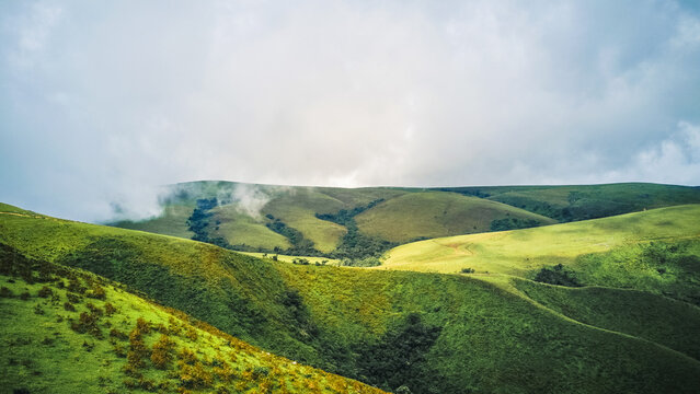 Aerial view of lush, undulating green hills bathed in dappled sunlight and soft shadows, with distant peaks shrouded in ethereal mist, Obudu, Cross River, Nigeria.