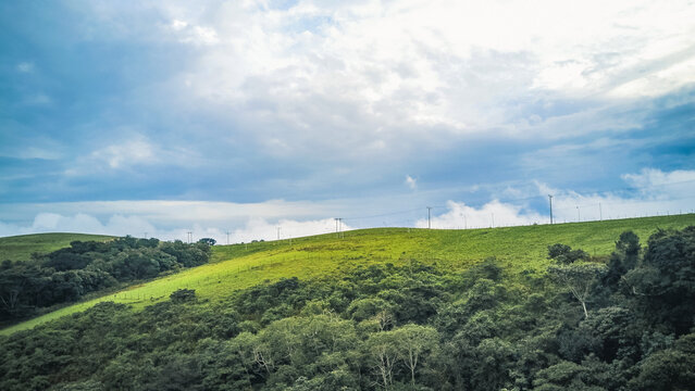 Aerial view of the vibrant lime green slopes meeting the dense, dark green forest under a dramatic sky of swirling blue and white clouds, Obudu, Cross River, Nigeria.