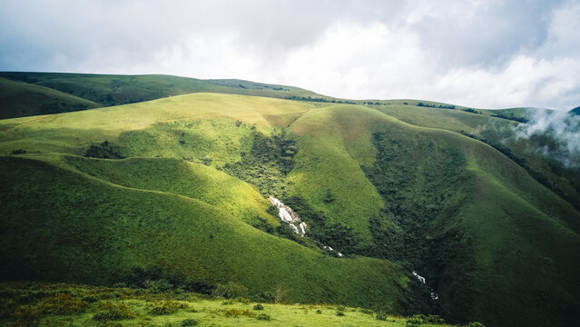Aerial view of rolling green hills cascade under a soft, diffused light, revealing pockets of deep shadow and a distant waterfall, Obudu, Cross River, Nigeria.
