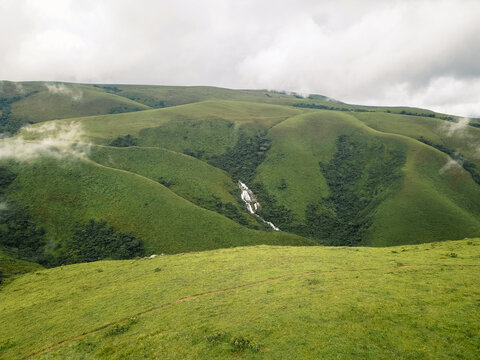 Aerial view of verdant rolling hills with a striking white waterfall and lingering mist beneath a grey sky, Obudu, Cross River, Nigeria.