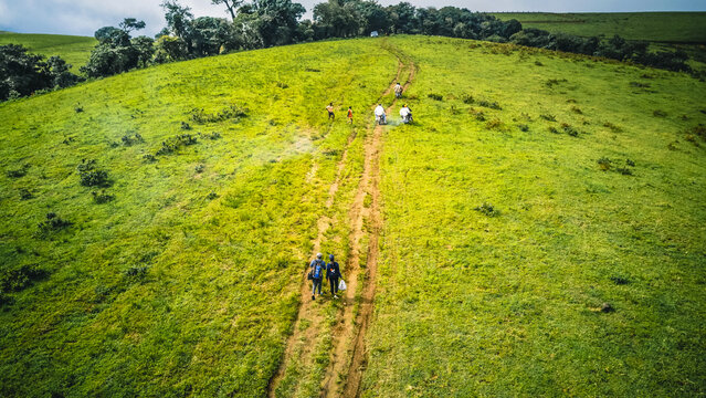 Aerial view of hikers trekking a winding dirt path across the vibrant, undulating green hills under a dramatic sky, Obudu, Cross River, Nigeria.