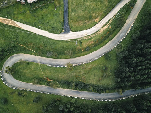 Aerial view of a winding asphalt road cutting through vibrant emerald green slopes and bordering a dark forest edge, Obudu, Cross River, Nigeria.
