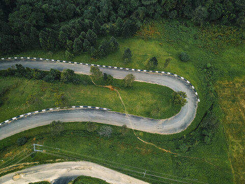 Aerial view of a winding asphalt road, its grey ribbon with checkered edges contrasting with vibrant emerald and olive green hillsides and dense forest, Obudu, Cross River, Nigeria.