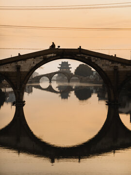 Aerial view of a lone figure perched atop an ancient arched bridge, its reflection mirroring a distant pagoda in the tranquil waters, Shaoxing, Zhejiang, China.
