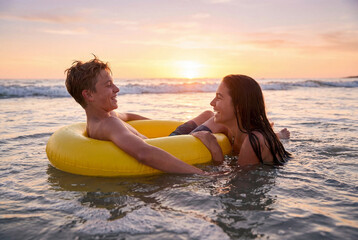 Obraz premium Teenage couple laughing in the ocean with a yellow inflatable ring at sunset. Concept of summer romance and carefree youth.