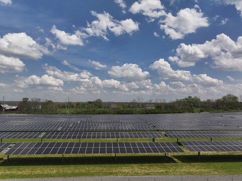 Aerial view of expansive solar panels reflecting the sky under fluffy clouds, contrasting against the green horizon, Lebanon, Ohio, United States.