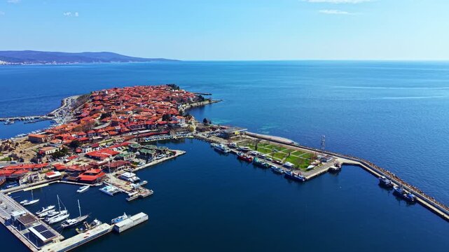 Nesebar Old Town appears from above with its old buildings, boats docked in the harbor, and the clear blue sea surrounding the town. A few clouds are in the sky.