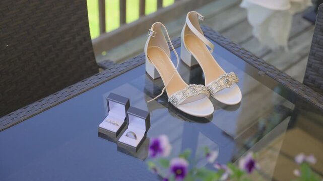 A high-angle shot showcasing elegant white bridal sandals with rhinestone embellishments alongside two wedding bands in open boxes, set on a reflective glass patio table.