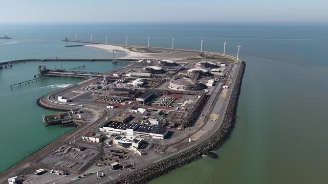 High angle drone shot slowly orbiting an expansive liquefied natural gas import terminal located on a coastal breakwater, highlighting the massive storage tanks and surrounding wind turbines