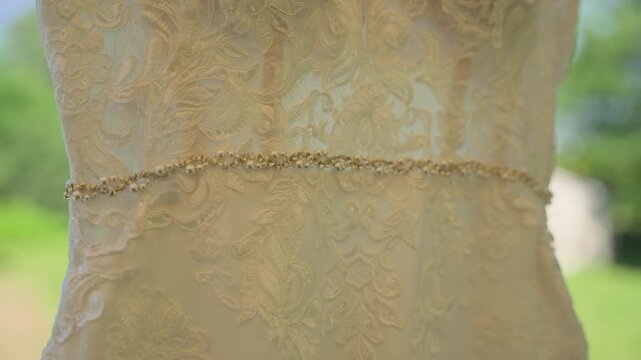 A stunning close-up showcasing the shimmering rhinestone belt and delicate floral lace of a wedding dress, with a soft-focus summer garden visible in the natural background light.