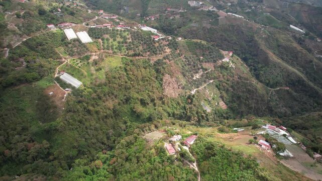 Drone view of mountainous agricultural landscape. Scene features winding dirt roads, scattered rural homes with red roofs, and dense tropical vegetation surrounding cultivated plots. Highland farming