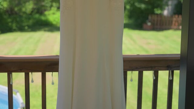 A vertical camera movement following the flowing white satin fabric of a bridal gown hanging from a wooden deck railing, with a blurry garden and pool in the background.