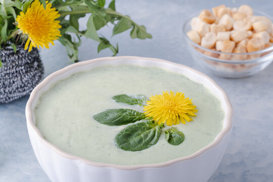 A bowl of fresh green soup sits on a blue background. A dandelion flower and leaves float on top. A bowl of crackers is placed nearby, ready to enjoy. Healthy and vegan food