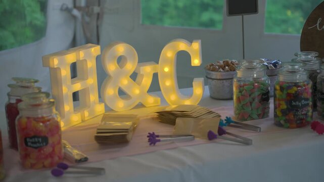 A charming wedding dessert table featuring bright marquee letters H and C and various glass jars filled with colorful candies and jelly beans at a festive reception.