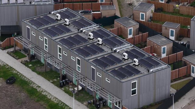 Aerial tracking shot of modern prefabricated apartment complex with flat roof covered in solar panels and heat pumps, highlighting sustainable, affordable urban housing solutions