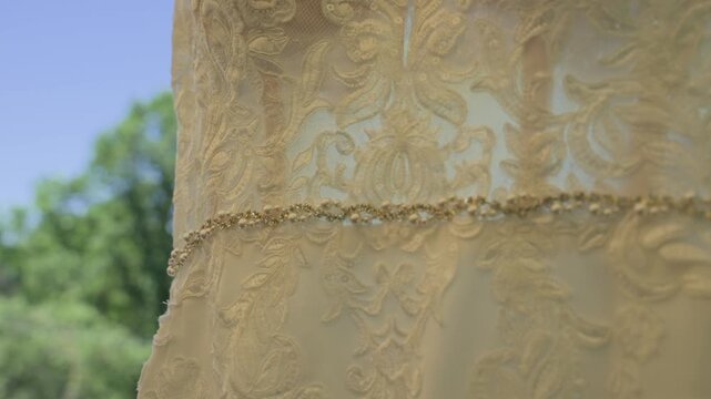 A detailed close-up video showcasing the shimmering rhinestone belt and delicate embroidery of a bridal dress, panning to reveal a soft-focus background of lush green trees and blue sky.