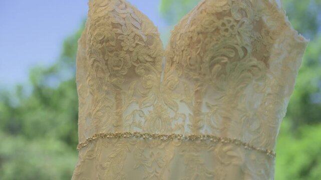 A detailed close-up of a white lace wedding gown featuring intricate floral embroidery and a sparkling rhinestone belt, hanging against a blurred background of green trees and blue sky.
