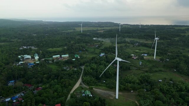 Aerial view of wind turbines rising above a rural village surrounded by lush greenery with coastal landscape in the background in Guimaras, Philippines during daytime