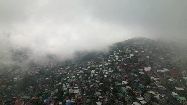 Aerial orbit circling a dense hillside community with colorful rooftops and fog drifting across Quirino Hill, Baguio City, Philippines