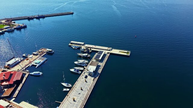 Visitors enjoy the view of the harbor in Nesebar Old Town. Boats are docked alongside piers while the water reflects the blue sky. The scene captures daily life in this seaside location.