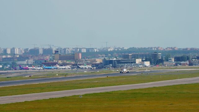 Commercial narrow body jet lifts off runway at busy international airport, air traffic control tower visible in background, multiple aircraft at gate, urban skyline beyond