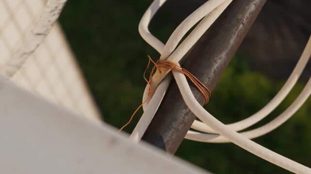A thick white cable is tied to a rusty metal pole using a thin piece of copper wire. The makeshift connection is outdoors with a soft blurred background of a fence and green foliage.