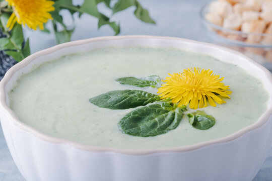 Close up of a bowl of fresh green soup sits on a blue background. A dandelion flower and leaves float on top. A bowl of crackers is placed nearby, ready to enjoy. Healthy and vegan food