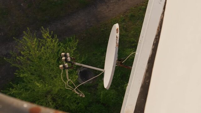 White satellite dish is mounted securely on the exterior wall of an urban apartment building. The high angle view looks down past the antenna hardware toward green trees and a manhole cover below.