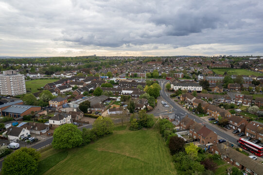 Aerial view of red double-decker bus winding through streets amidst verdant trees and rooftops under a cloudy sky, Slade green, United Kingdom.