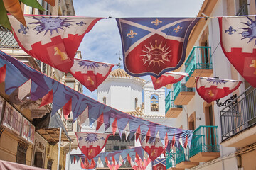 Multiple heraldic banners with sun face emblems, fleur de lis and castle motifs layered across narrow street with bunting and church facade in background in Santa Fe, Spain, traditional celebration © Goodwoon Mallorca