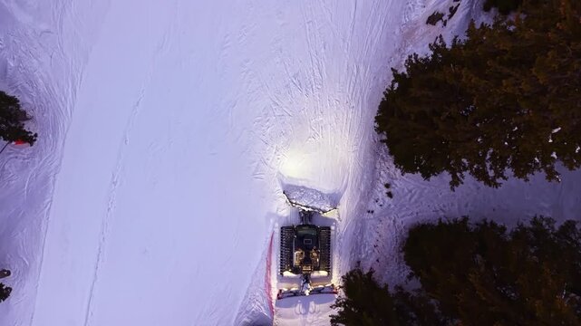 A snow groomer works on a ski trail at night in a winter landscape. Trees surround the area, and the machine lights up the snow as it moves along the path.