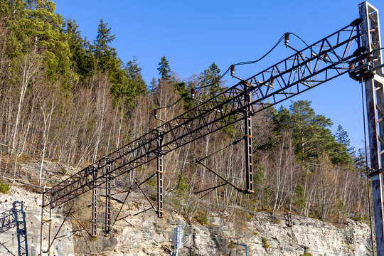 A modern catenary system spans across a railway track nestled against a rugged rocky hillside and lush forest, illustrating efficient transportation infrastructure and precise engineering for travel