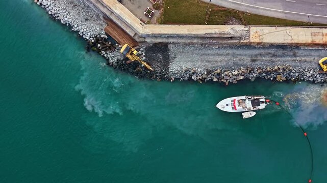 Workers use machines on the shore while a boat operates in the water. Construction equipment is visible along the coastline. This is a busy work site.