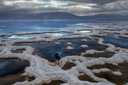 Aerial view of a lone figure exploring the surreal, brilliant white salt formations contrasting with the deep blue waters of the Dead Sea under a dramatic sky, Dead Sea, Israel.